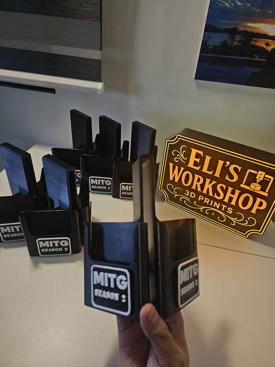 Several black 3D printed desk card holders with a raised logo badge on the front, arranged on a table beside a lit Eli's Workshop sign.