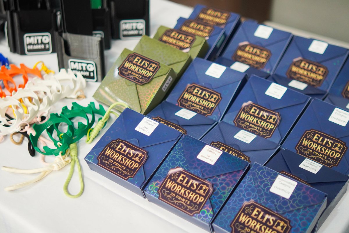 Rows of branded Eli's 3D Workshop gift boxes laid out on a table with category labels, alongside 3D printed keyrings and desk holders, prepared as corporate giveaways.
